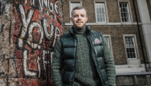 © IWM and shows Russell Tovey standing in front of a section of the Berlin Wall at IWM London, for Series 2 Episode 1 of Conflict of Interest on the Berlin Wall.