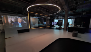 A photograph of the exhibition shows a large space with a circular light overheard, and a projection of Al Jazeera news on the wall