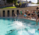 A photograph captures a group of swimmers mid-leap into an outdoor pool