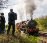 A steam engine travels along tracks as onlookers watch from an embankment
